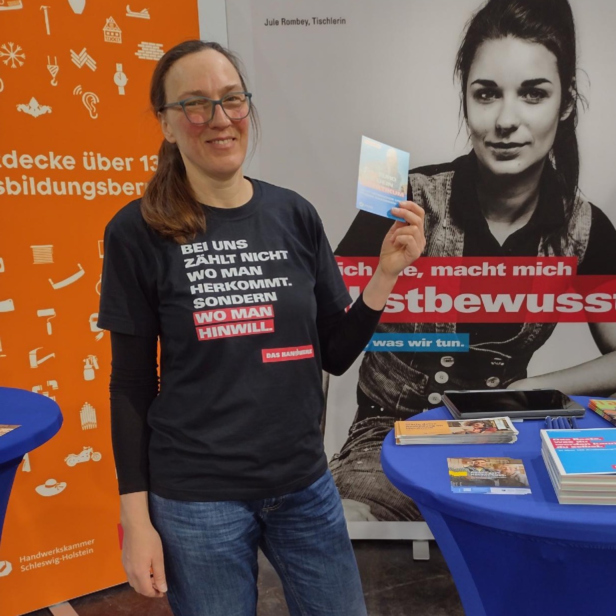 KI generiert: Eine Frau steht vor einem Informationsstand und hält ein Kärtchen, neben einem Plakat und Broschüren, die Berufe und Selbstbewusstsein thematisieren.