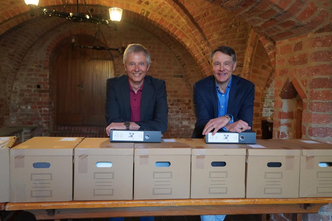 2 Männer stehen hinter zahlreichen Kartons mit Ordnern.