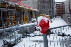 KI generiert: Ein schneebedeckter Bauhelm hängt an einem Bauzaun vor einer verschneiten Baustelle.