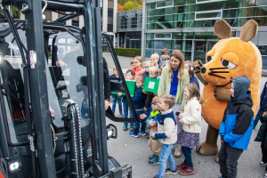 KI generiert: Eine Gruppe von Kindern steht neben einer Person in einem großen Kostüm in Form einer orangefarbenen Maus, während sie einen Gabelstapler betrachten.