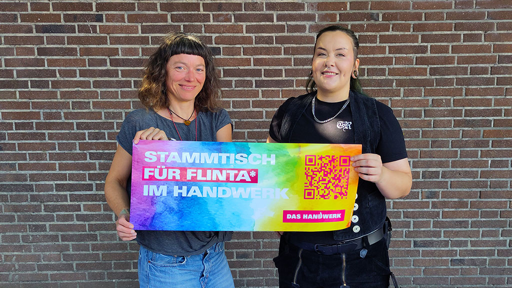 KI generiert: Zwei Personen halten ein buntes Schild mit der Aufschrift "Stammtisch für FLINTA* im Handwerk" vor einer Backsteinmauer.