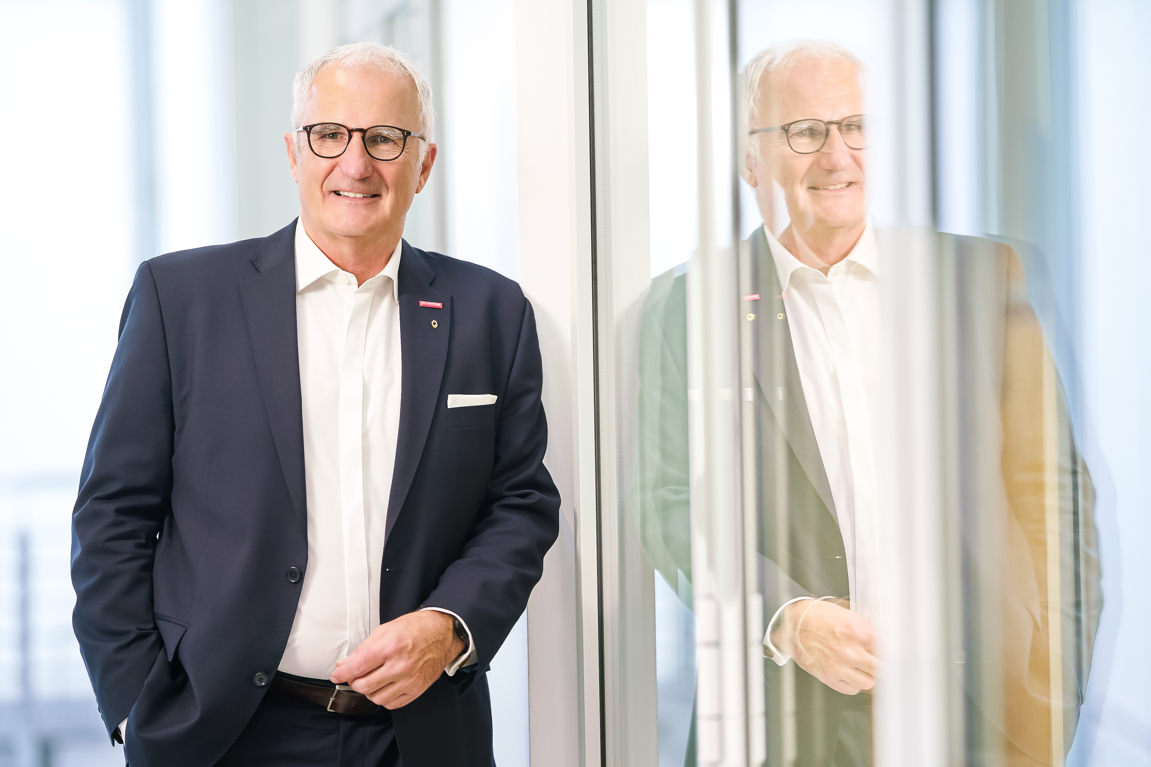 KI generiert: Ein Mann in einem Anzug steht lächelnd neben einer Glaswand, in der er sich spiegelt.