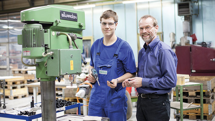 Azubi und Ausbilder stehen vor einer Maschine in einer Werkstatt