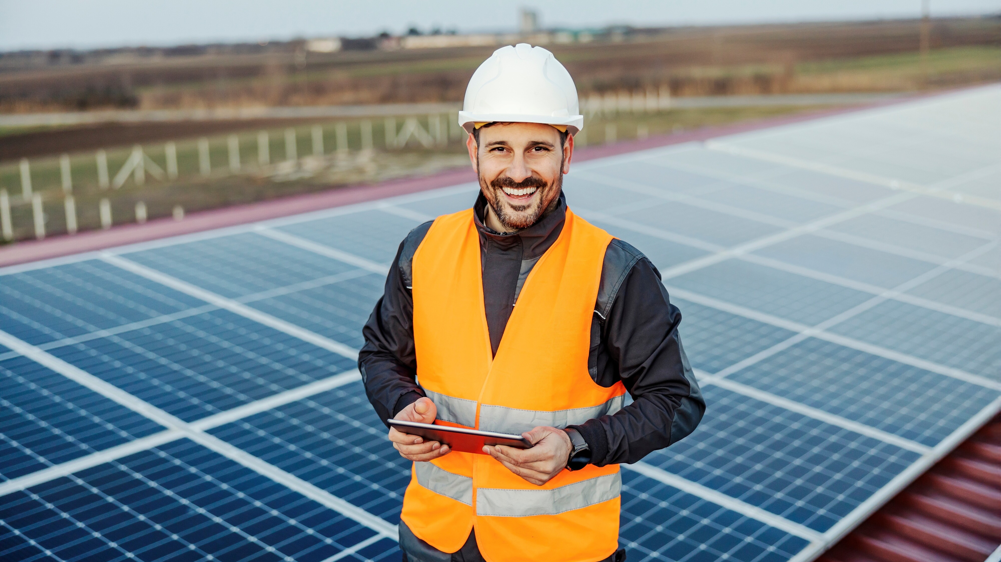 KI generiert: Ein lächelnder Mann in Sicherheitskleidung und Helm steht vor einer Reihe von Solarpanels und hält ein Tablet.